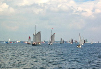 Sailboats and yachts on the water, viewed from a houseboat, with a cloudy sky and harbor in the distance.