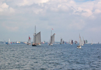 Zeilboten en jachten op het water, gezien vanaf een woonboot, met haven en bewolkte lucht op de achtergrond.
