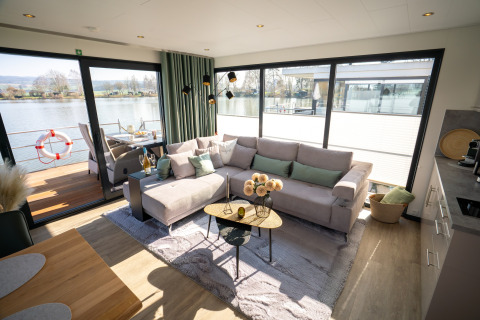 Salón en una casa flotante en Höxter, con sofá grande, decoración moderna y vistas al río desde las ventanas.