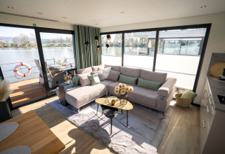 Stue med store vinduer på en husbåd i Höxter, komfortabel sofa og udsigt over floden udenfor.