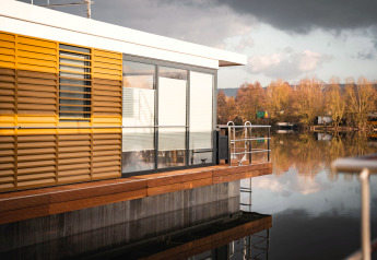 Moderne woonboot in Höxter met gele luiken en grote ramen, omringd door kalm water en natuur.