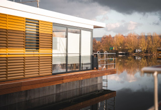 Houseboat moderno a Höxter con persiane gialle e grandi vetrate sul lago calmo e alberi autunnali.