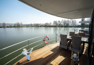 Terraza de una casa flotante en Höxter con vista al río, conjunto de comedor y sillas al aire libre.