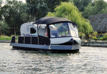 Houseboat named 'Watercamper deluxe' cruises on a river in Germany with lush trees and cottages nearby.