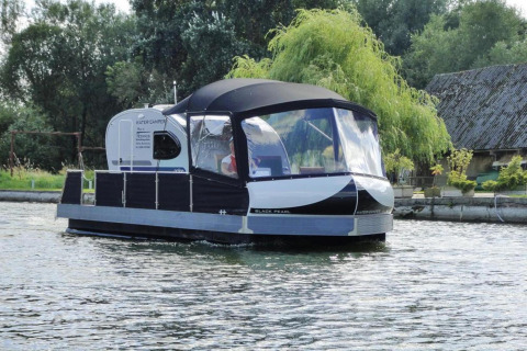 Woonboot genaamd 'Watercamper deluxe' vaart op een rivier in Duitsland met bomen en huisjes op de achtergrond.