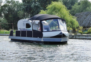 Woonboot genaamd 'Watercamper deluxe' vaart op een rivier in Duitsland met bomen en huisjes op de achtergrond.