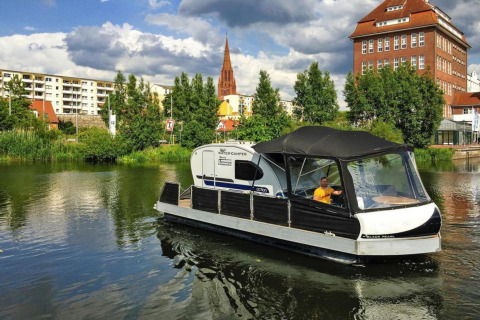 Een man vaart met een woonboot 'Watercamper deluxe' over een rivier in Duitsland, met stad op de achtergrond.