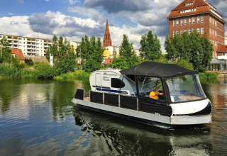 Een man vaart met een woonboot 'Watercamper deluxe' over een rivier in Duitsland, met stad op de achtergrond.