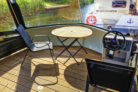 Zonnige zithoek met tafel, stoelen en stuurwiel op een Watercamper deluxe woonboot in Duitsland.
