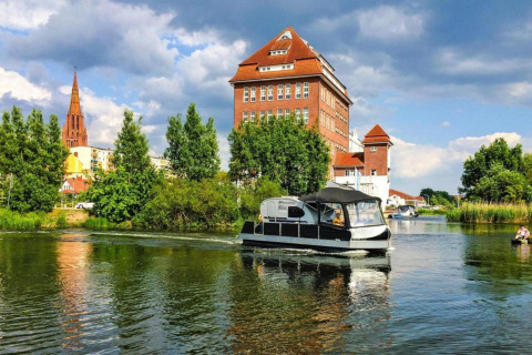 Watercamper deluxe houseboat vaart op een rivier met een kerk en historische gebouwen op de achtergrond.