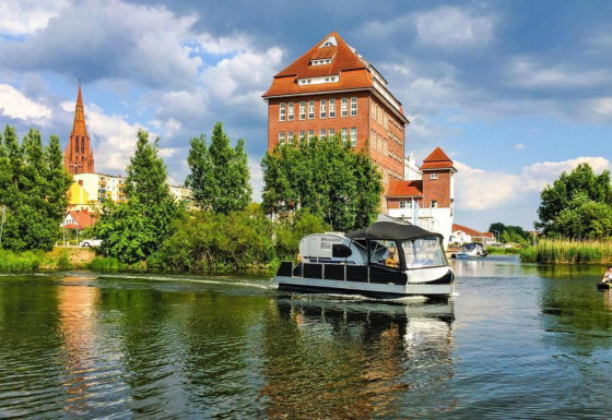 Watercamper deluxe houseboat vaart op een rivier met een kerk en historische gebouwen op de achtergrond.