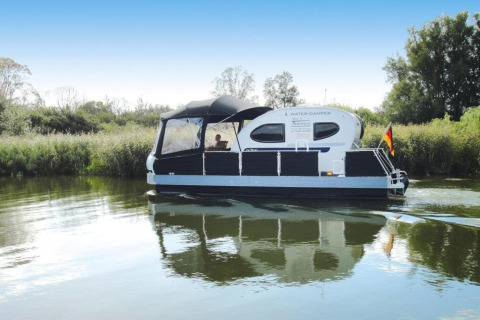 Woonboot met de naam Watercamper deluxe vaart op een rivier in Duitsland, omringd door groene natuur.