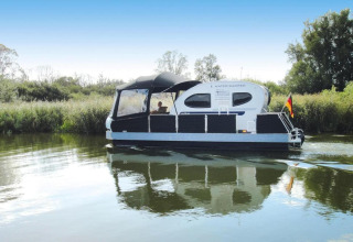 Woonboot met de naam Watercamper deluxe vaart op een rivier in Duitsland, omringd door groene natuur.