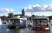 Casa flotante en el puerto de Ribnitz-Damgarten, Alemania, con la ciudad y torre de iglesia al fondo.