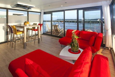 Sala moderna en una casa flotante con sofás rojos, mesa de comedor y vistas al agua en Ribnitz-Damgarten, Alemania.
