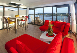 Modernes Wohnzimmer auf einem Hausboot mit roten Sofas, Esstisch und Wasserblick in Ribnitz-Damgarten, Deutschland.