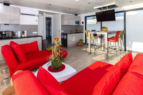 Casa flotante moderna con sofás rojos, cocina luminosa y sillas de colores, en Blue Active, Alemania.