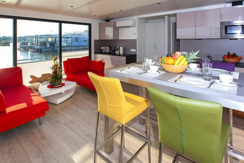 Salon moderne et cuisine sur une péniche avec chaises et canapés colorés, vue sur l’eau en Allemagne.