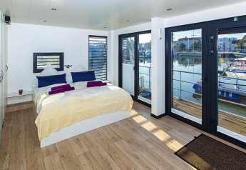 Dormitorio en casa flotante con grandes ventanales y vistas al puerto en Ribnitz-Damgarten, Alemania.