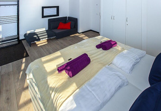 Dormitorio moderno en una casa flotante con cama doble, toallas moradas, sofá y armarios blancos.