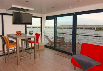 Intérieur d'une péniche moderne avec chaises colorées, table, et vue sur l'eau par de grandes fenêtres.