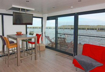 Interior de una casa flotante moderna con sillas coloridas y vista al agua a través de ventanas grandes.