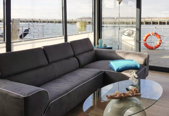 Wohnbereich im Hausboot 'Leni' mit grauem Sofa, Glastisch und Blick aufs Wasser in Ribnitz-Damgarten.