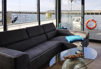 Sala de estar en la casa flotante 'Leni' con sofá gris, mesa de vidrio y vista al agua en Alemania.