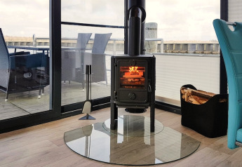 Modern wood stove on a glass plate inside a cozy houseboat living room, overlooking deck and water.