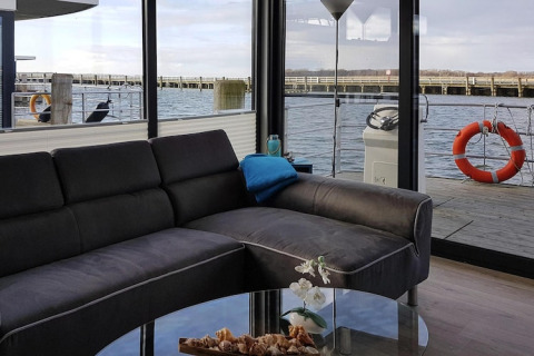 Sala moderna en una casa flotante con sofá oscuro y grandes ventanales hacia el agua, Ribnitz-Damgarten.