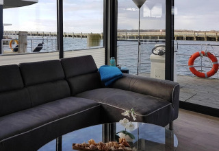 Salon moderne sur une péniche avec canapé foncé et vue sur l’eau depuis de grandes baies vitrées.