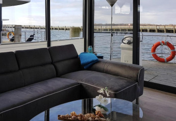 Modern living area on a houseboat with a dark sofa and glass walls overlooking water, Ribnitz-Damgarten.