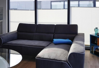 Moderne stue på husbåd med mørkegrå sofa, blå pude, store vinduer og bogreol i hjørnet.