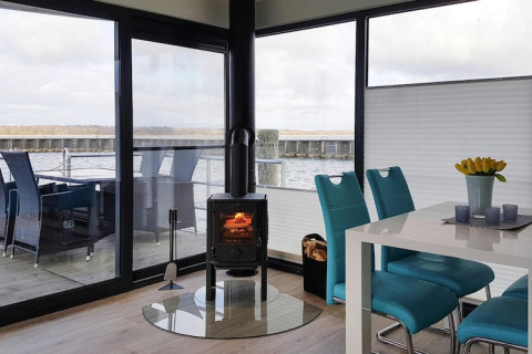 Moderne husbåd med panoramavinduer, træpilleovn, spisebord og blå stole med udsigt til sø og terrasse.