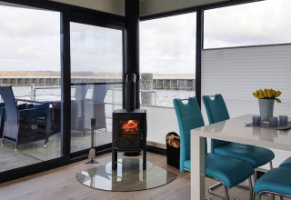Modernes Hausboot mit Panoramafenstern, Ofen, Esstisch und blauen Stühlen mit Seeblick und Terrasse.