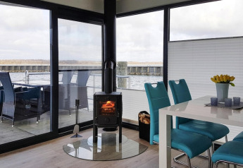 Intérieur de péniche moderne avec baies vitrées, poêle, chaises turquoise, table et vue sur le lac.