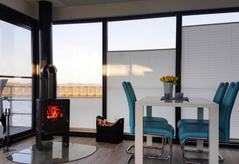 Interior acogedor de casa flotante con estufa, mesa de comedor, sillas azules y vista al agua en Ribnitz-Damgarten.