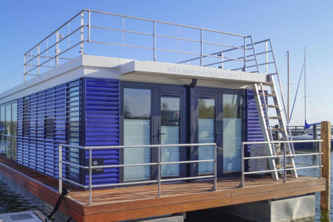 Casa flotante moderna con contraventanas azules y terraza en el puerto de Ribnitz-Damgarten, Alemania.