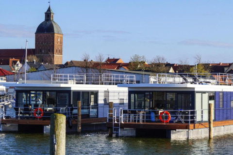 Casa flotante «Floating House Luisa» en Ribnitz-Damgarten, Alemania, con iglesia y ciudad al fondo.