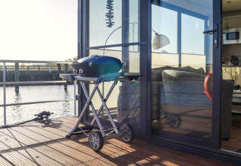 Ein Grill steht auf dem Deck eines Hausboots am Wasser, mit Blick in das gemütliche Wohnzimmer durch die Glastüren.