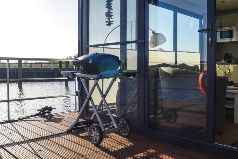 A grill is placed on the deck of a houseboat by the water, with a cozy living room seen through glass doors.