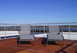Dos tumbonas en la terraza de una casa flotante con vistas al agua y al cielo azul en Alemania.