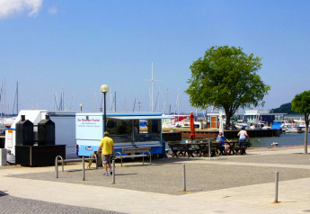 Hafenbereich mit Imbisswagen, Sitzbänken, Menschen und Segelbooten in Ribnitz-Damgarten, Deutschland.