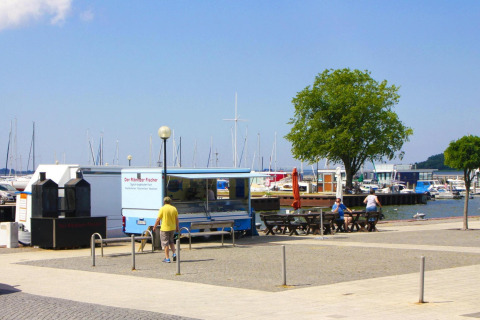 Vista del puerto con camioneta de comida, bancos, personas y veleros en Ribnitz-Damgarten, Alemania.