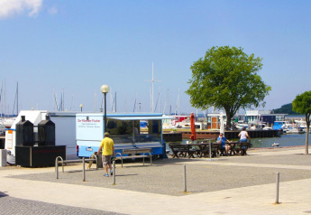 Vista del puerto con camioneta de comida, bancos, personas y veleros en Ribnitz-Damgarten, Alemania.