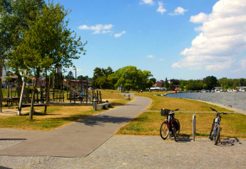 Uferweg in Ribnitz-Damgarten mit Fahrrädern, Spielplatz, Bäumen und Hausbooten bei sonnigem Wetter.