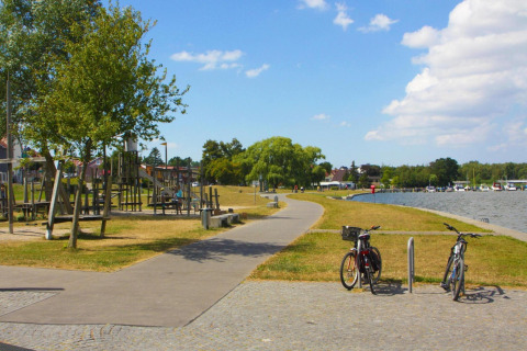 Uferweg in Ribnitz-Damgarten mit Fahrrädern, Spielplatz, Bäumen und Hausbooten bei sonnigem Wetter.