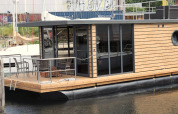Moderne woonboot ‘Leni’ met houten bekleding en glazen deuren aangemeerd in Duitsland, terras zichtbaar.