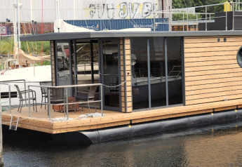Moderne husbåd 'Leni' med træpaneler og glasdøre ved en havn i Tyskland. Udsigt over dækket.