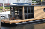 Péniche moderne ‘Leni’ avec bardage bois et portes vitrées amarrée en Allemagne, terrasse extérieure visible.